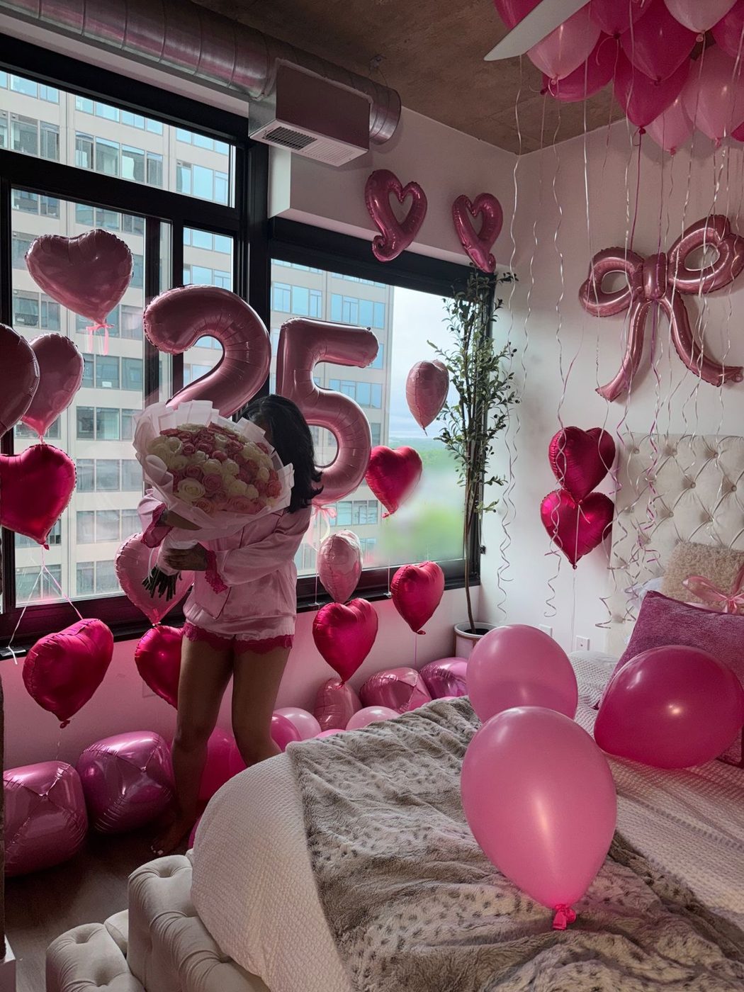 Birthday guest holding a rose bouquet in a room filled with pink heart balloons and 25 number balloons