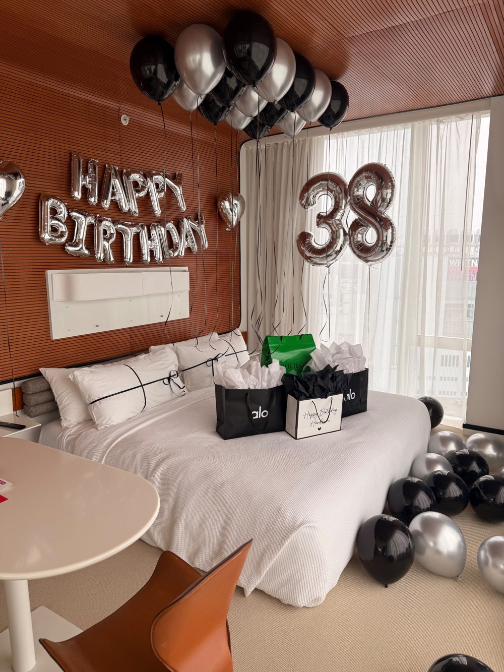 Hotel suite styled with black and silver balloons, a Happy Birthday banner, 38 number balloons and gift bags on the bed